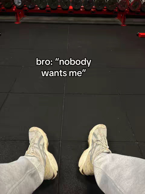 The image shows a person's legs and sneakers in a gym setting. The person is wearing light grey sweatpants and beige sneakers. The background features gym equipment, including a weight rack. Text overlay: bro: “nobody wants me”.