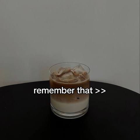 A glass of iced coffee sits on a black surface, likely a table, with a plain off-white background. The coffee has two distinct layers, a lighter shade at the bottom and a darker brown at the top, filled with ice cubes. The glass is positioned slightly off-center to the right.