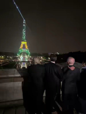A nighttime shot of the Eiffel Tower in Paris, illuminated in green, yellow, and blue lights. Standing in the foreground are several people with their backs to the camera, looking towards the tower. The composition is from an elevated vantage point.