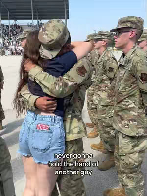 The image shows a couple embracing in an outdoor location, potentially a military base. The woman, in a blue shirt and jean shorts, is hugging a man in military uniform. Other uniformed figures stand in formation in the background, with bleachers filled with observers. The warm, sunny weather provides a backdrop for the intimate embrace, suggesting a reunion or emotional moment with the text overlayed at the bottom.
