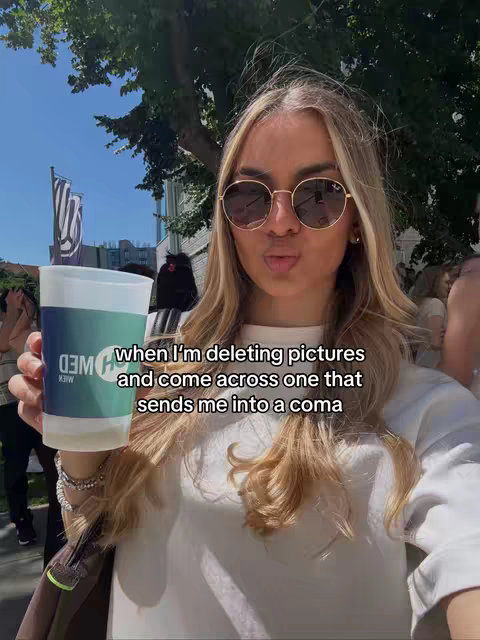The image showcases a young woman with long, wavy blonde hair wearing round sunglasses and a white t-shirt. She's outdoors on a sunny day, with a crowd in the background.  She holds a cup in front of her with a design on it.