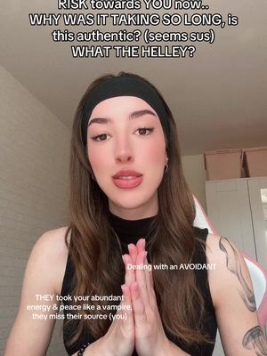 A woman is centered in the frame, her hands clasped in front of her chest in a prayer-like gesture. Above her head, overlaid text asks, 'RISK towards YOU now.. WHY WAS IT TAKING SO LONG, is this authentic? (seems sus) WHAT THE HELLEY?' Below, additional text reads, 'Dealing with an AVOIDANT' and 'THEY took your abundant energy & peace like a vampire, they miss their source (you).' The woman has brown hair and is wearing a black headband and top. The background is a blurred interior with a white wall and chair.