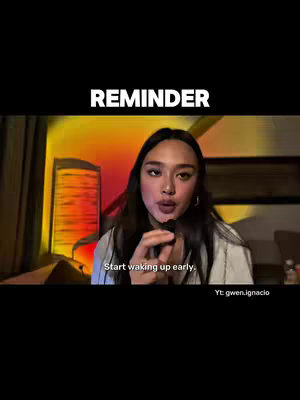 A woman with long dark hair is holding a microphone and speaking. Above her head, the word "REMINDER" is written in large, white letters. Below her, text reads, "Start waking up early." The background is dimly lit, with a blurred view of a building and some light sources.