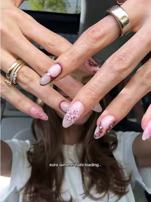 A close-up shot of a person's hands with elaborately decorated nails. The hands are crossed, and the focus is on various nail designs. The nails have a light pink base with detailed art, including floral patterns, and are accented with rhinestones and a dark red border.