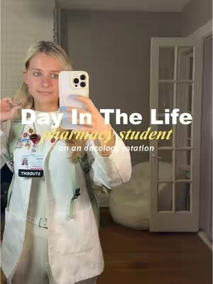 A young woman in a white lab coat is holding a phone up to take a selfie. She has blonde hair, and is wearing a lanyard with a photo ID, and a name tag that reads "THBOUTZ". The text overlay reads 'Day In The Life pharmacy student on an oncology rotation'. Behind her is a mirror and other doors.