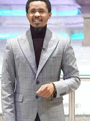 A man in a grey plaid suit and a burgundy turtleneck is the central focus. The background includes a series of light blue illuminated panels. The man is smiling with a friendly expression. He has a black round wig, he's wearing a bracelet, and he has his right hand on his side, his face expressing happiness and intelligence.