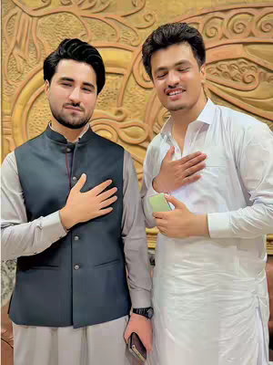 Two men are posing with their hands over their hearts. The man on the left is wearing a black vest and a light grey shirt. The man on the right is wearing a white outfit and holding a phone. Behind them is a golden wall with a carved design.