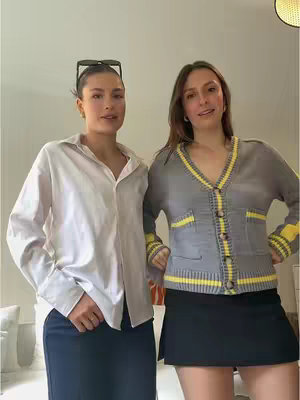 Two young women stand together indoors, posing slightly towards the camera. The woman on the left wears a white button-down shirt and dark pants with sunglasses on top of her head, and the other wears a gray cardigan with yellow accents over a black skirt. The background is plain, with neutral-colored walls and a glimpse of a ceiling light fixtures.