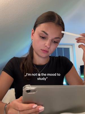 A young woman with brown hair in a ponytail is looking down pensively while holding a stylus to her forehead. She is wearing a black t-shirt. The text overlay reads, "I'm not in the mood to study" and is located on the bottom left of the frame. In the background is a simple domestic interior with a window with a blue-ish light and a ceiling.