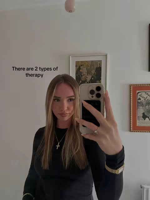 A woman is taking a selfie in front of a white wall, holding her phone up to her face. The text overlay on the screen says 'There are 2 types of therapy'. She wears a black long sleeve shirt, a cross necklace, a gold bracelet watch and a ring on her finger. There are framed pictures hung on the wall in the background.