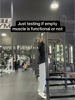 A man is seen in a gym setting, appearing to perform a muscle-up on a pull-up bar. He is in the midst of the motion, with his body angled up. The setting is well-lit, and a text overlay is present at the top, set within a black rectangle and indicating the man is demonstrating his ability. In the background, additional gym equipment and other individuals exercising are visible.