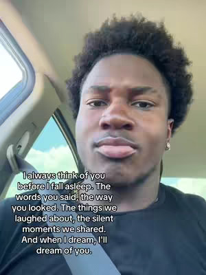 A selfie-style shot of a young Black man inside a car. The man has short, dark, curly hair and is wearing a dark-colored t-shirt. He has a slightly puckered expression, as if he's about to kiss or is deep in thought. The background suggests the interior of a vehicle, with a glimpse of the car's interior visible.
