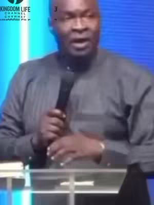 A medium shot captures a man with short, dark hair and a gray button-down shirt speaking into a microphone. He stands behind a clear podium in a room with a teal-colored background. The video shows his face. At the top left corner is a white text overlay.