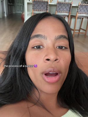 A selfie-style video cover. The creator is a woman with long, dark hair, facing the camera with an open mouth, possibly mid-speech. The background includes a glimpse of a light-colored room with chairs. Text overlay on the image reads 'the process of a cancer' with a purple emoji.