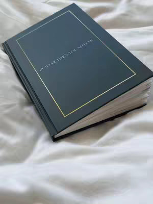 A book with a dark teal cover and gold border lies open on a white bed sheet. The book's title, "READ ME WHEN YOU NEED ME" is visible on the cover. The scene is lit with soft, natural light.