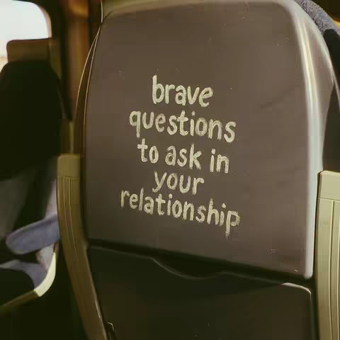 The image shows the back of a chair in what looks like a public setting, with text written on the chair back. The chair back is a dark gray color, with the text written in a lighter silver or white color and is slightly worn. The overall color palette is muted and neutral, with focus on the text in the image.