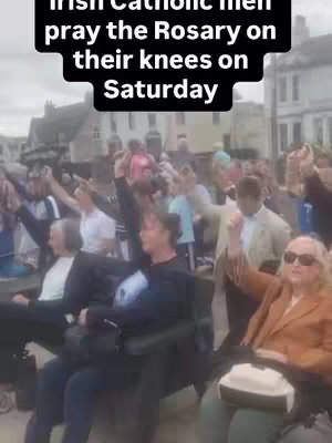 A group of people, mostly men, are shown outdoors. Many are kneeling with their hands raised, holding what appear to be prayer beads, directed upwards, signifying prayer. The background suggests an urban setting with buildings visible. An overlay on top reads: Irish Catholic men pray the Rosary on their knees on Saturday.