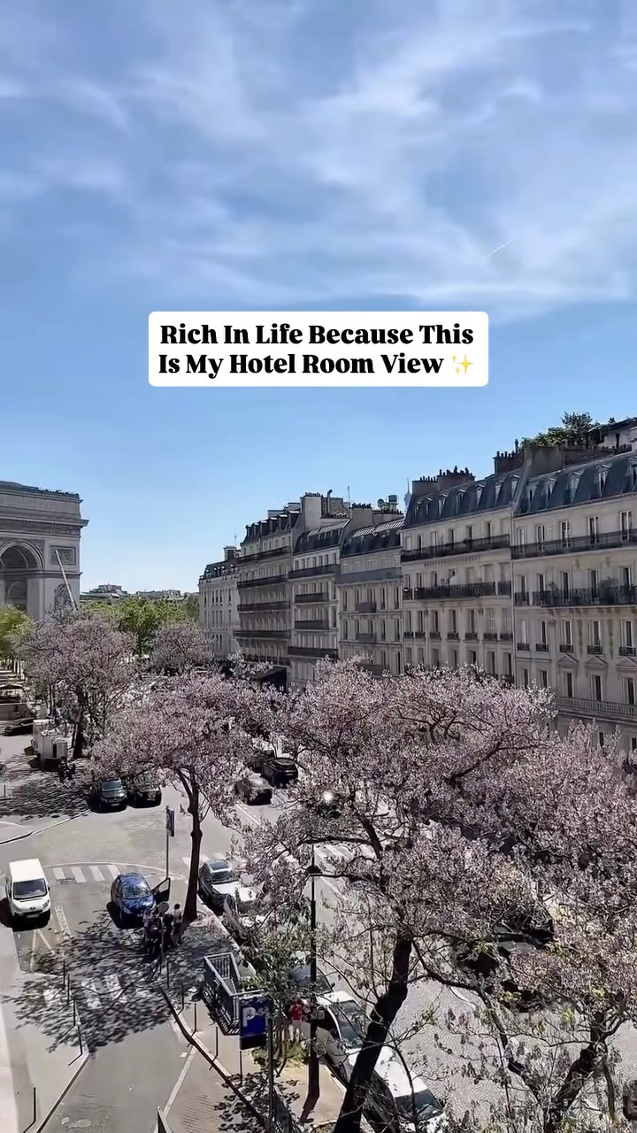 An overhead shot of a Parisian street lined with buildings. The Arc de Triomphe is visible on the left edge. The foreground features pink flowering trees. A text overlay reads, 'Rich In Life Because This Is My Hotel Room View ✨'.