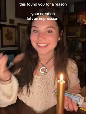 A smiling young woman with long dark hair is holding a lit gold candle and a deck of tarot cards. She is wearing a white lace top and a silver necklace with a spiral pendant. Decorative items are present in the background.