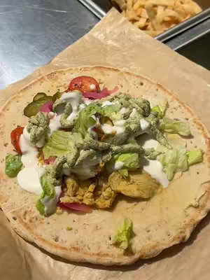 The image is a top-down shot of a pita wrap, filled with colorful ingredients. The wrap is open and the filling is visible, which includes a base of yellow-toned food, possibly chicken shawarma, topped with white sauce, green lettuce and herbs, red tomatoes, and pickled cucumbers. The colors are vibrant and appetizing, with a warm, natural light. The wrap sits on a piece of brown paper, and next to it there is a metal surface, the background includes a tray of food that seem to be french fries.