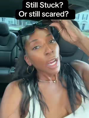 A woman with long black hair sits in a car. Sunglasses are placed on her head. The background is blurred with outside.