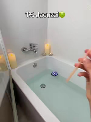 A bathroom scene with a white bathtub filled with water. Two lit candles and other decorative elements are visible. A hand holds a stick, likely for testing or indicating the water's state; a hook's text is overlaid on the top left.