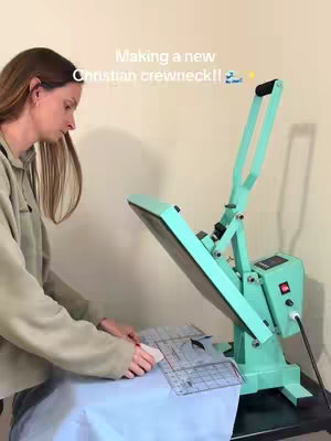 A woman is working on a garment using a heat press machine. The machine is primarily teal in color. The woman is wearing a beige hoodie, and the setting appears to be an indoor space.