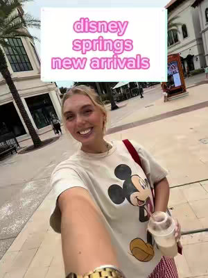 A selfie-style image with a young woman smiling and holding a drink, set against the backdrop of Disney Springs. She is wearing a Mickey Mouse t-shirt and a gold watch. A text overlay states "disney springs new arrivals". The background includes shops, palm trees, and other people.