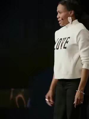 A woman stands in a dimly lit setting, slightly turned to her side, wearing a white sweater with the word 'LOVE' printed on it and black pants.