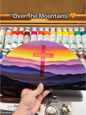 The image shows a hand holding up an oil painting. The painting depicts a landscape with low, layered purple and grey mountains beneath a gradient sky of orange, yellow, and a hint of purple; a cross is centered in the sky. The painting is semi-circular and displayed against a background of artist's supplies, including paint tubes. The hand holding the artwork is visible in the foreground.