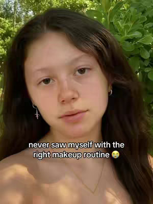 A young woman with long brown hair looks directly at the camera. The background is blurred green foliage, suggesting an outdoor setting. Text is overlaid at the bottom, partially obscuring her chest and a gold necklace.