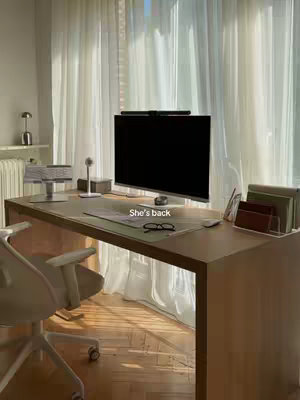 An office interior with a large wooden desk, a computer, and sheer curtains. The desk has a monitor with a blank screen, keyboard, and other office supplies. The lighting is soft and diffused, suggesting a window. The image has a neutral color palette with beige, white, and brown tones.