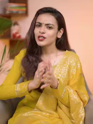 A woman with long brown hair and a yellow outfit is shown in a close-up shot, seated with her hands clasped together in front of her. She has a small bindi on her forehead and red lipstick. The background is blurred, suggesting an indoor setting with warm lighting.