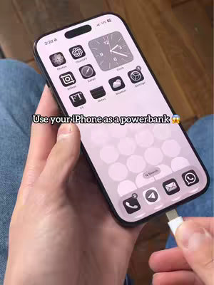 A person's hands holding an iPhone with the screen on, showing an app interface. There is a white USB-C cable plugged into the bottom of the iPhone. The iPhone has a light-colored background with various app icons, and the scene takes place on a person's lap, likely a jean.