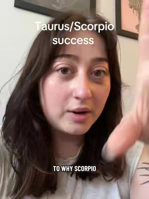 A young woman with dark hair is centered in a selfie view. The top of the image features text overlays: "Taurus/Scorpio success" above, and "TO WHY SCORPIO" at the bottom. Her expression is a mix of curiosity and engagement, and she seems to be pointing off-screen with her right hand. The background is blurred, suggesting an indoor setting with framed artwork. The overall composition is close-up and personal, enhancing its direct engagement.