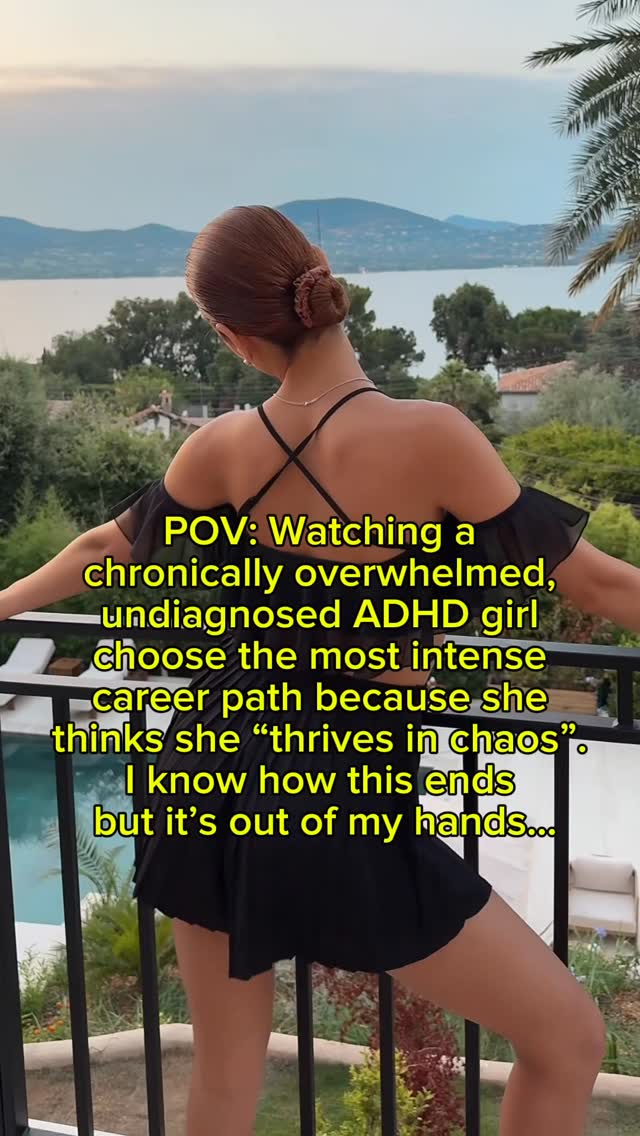 A woman with auburn hair styled in a bun stands on a balcony overlooking a scenic landscape with a lake and mountains. The woman is wearing a black dress. Text overlays in yellow cover some portion of the frame.