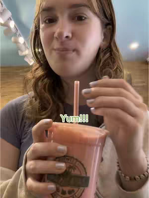 A young woman with long, wavy brown hair is holding a pink smoothie in a clear plastic cup. She is wearing a gray shirt and a white sweater. The background includes a wooden wall and a section of a ceiling with lights. The overlay features the text "Yum!!!".