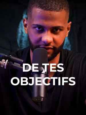A man with a beard and short hair looks directly at the camera, holding a microphone. The words "DE TES OBJECTIFS" are overlaid in white, bold font near the bottom of the image. The background is dark, with a hint of a blurry figure.