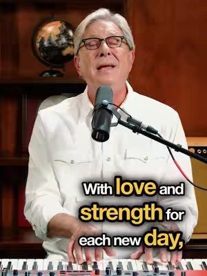 An older man with glasses is playing a white and black keyboard while singing. Below him, a text overlay says, 'With love and strength for each new day'. The background has a microphone, globe, and a wooden cabinet. The lighting is warm.
