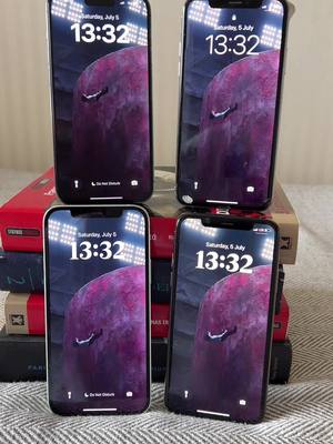Four smartphones are stacked in a square, each displaying a similar wallpaper of a large, abstract form with purple and gray hues. The background consists of books stacked on top of each other with the screens facing the viewer.