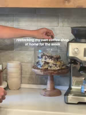 View shows a kitchen counter with a coffee machine on the right and stacks of white cups on the left. A wooden cake stand holding cake is in the center with a woman's arm reaching over to the food. Text overlaid says, "restocking my own coffee shop at home for the week."