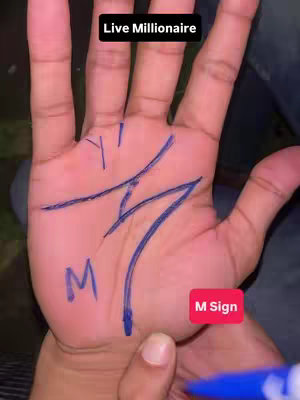 A close-up shot of a hand with palmistry markings. The palm is light-skinned and the focus is on the lines drawn in blue ink, forming an "M" shape. Above the hand is the text "Live Millionaire" in a black box, and at the bottom right, there's a red rectangular tag that says "M Sign". The background is blurred.