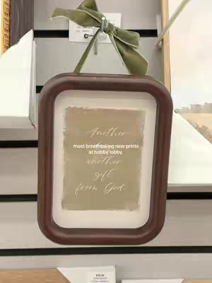 A framed piece of art hanging in a store. The frame is dark brown with a rounded edge. Inside is a light beige rectangle with text that reads 'Another most breathtaking new prints at hobby lobby another gift from God'. A light green bow decorates the top of the frame.