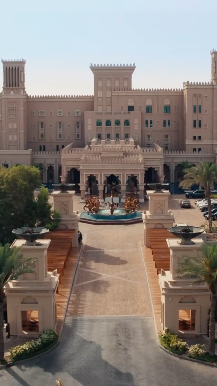 An aerial view of a large, ornate building with a symmetrical facade. The building is a light beige color with multiple levels and architectural details, including towers and arched entryways. A long pathway leads up to the entrance, flanked by decorative elements and palm trees, suggesting a luxurious or resort-like setting.