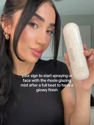 A woman with long dark hair and makeup holds a white Rhode skin glazing mist bottle against her face. Text overlays the image, and the composition is set against a neutral background.