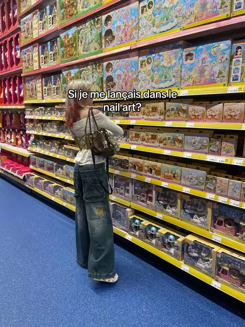A person stands facing away from the camera, in a store aisle filled with colorful boxes and toys. The floor is a bright blue, and the overhead lighting reflects in the aisles. The person is wearing loose-fitting jeans, and a top with a small purse.