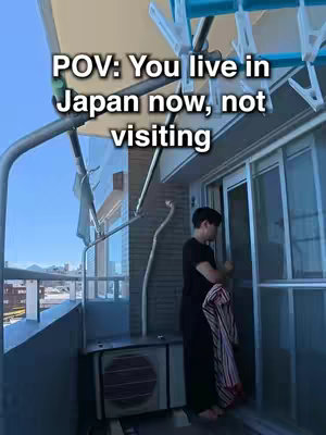 The image shows a person on a balcony in Japan. The person wearing a black shirt and dark pants. They are reaching for a door. The image has text overlay in white font stating 'POV: You live in Japan now, not visiting'. The lighting is bright and the sky is blue.