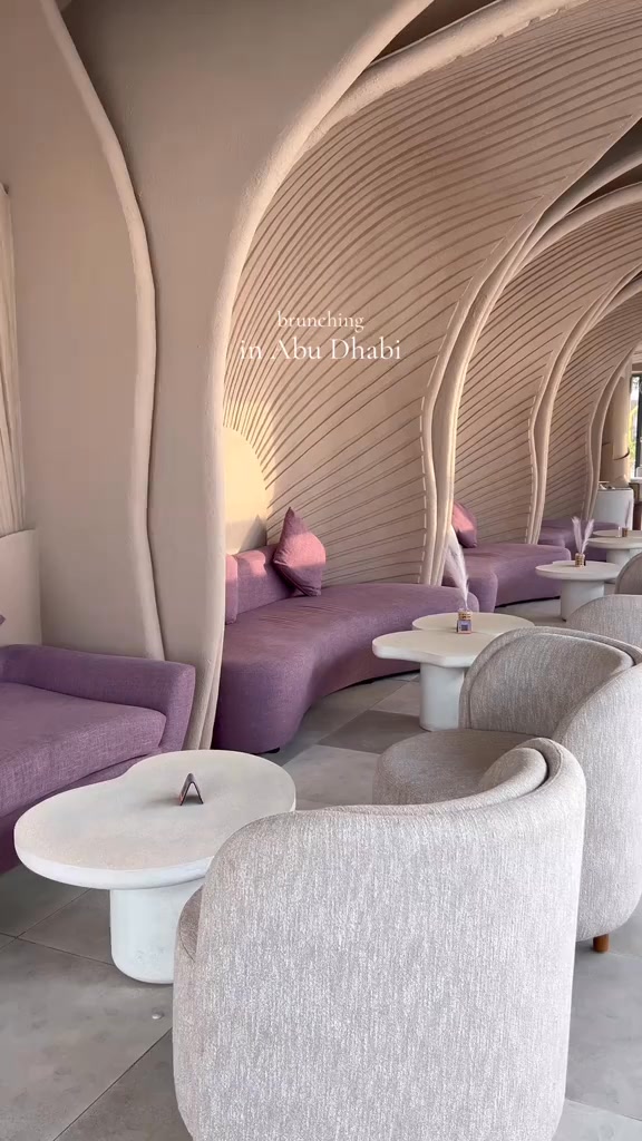 An interior shot of a modern restaurant lounge area. Serpentine purple couches and white round tables are arranged along the right side, while light gray armchairs and similar tables face them. The ceiling has a distinctive, abstract design with wave-like panels. The lighting is soft and warm with natural light coming from the left. In the background, there's a glimpse of the outside with more seating.