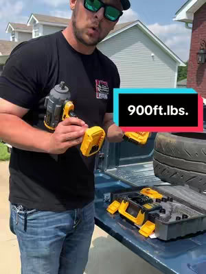 A man in a black t-shirt and sunglasses holds a yellow and black impact wrench in front of a truck bed. A text overlay reads "900 ft. lbs." in a blue box. A second tool kit and a tire are in the background.