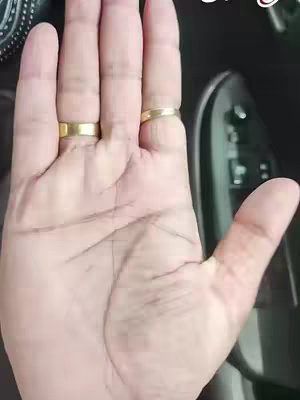 The image shows a close-up of a person's open hand, viewed from above. The palm and fingers are visible, with visible lines and skin texture. Two gold rings adorn the ring and middle fingers. The background is blurred, suggesting the image was taken inside a vehicle.